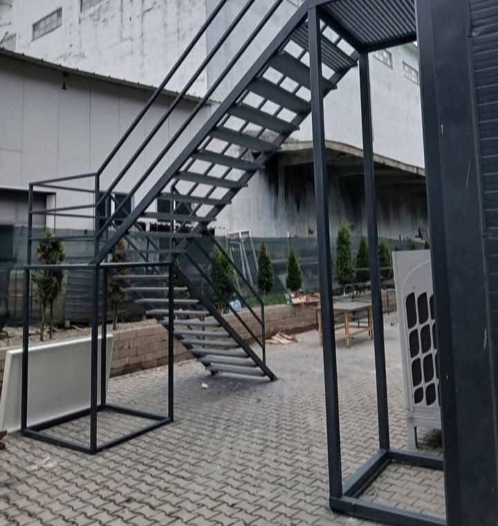 Scară metalică gri antracit, cu podest pe cadru propriu și balustradă pe ambele părți, într-o curte industrială