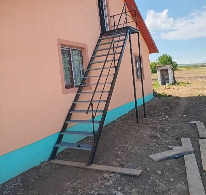 Scară metalică exterioară vopsită în negru, cu trepte gri și podest pe doi stâlpi, montată pe peretele unei case până la ușa mansardei