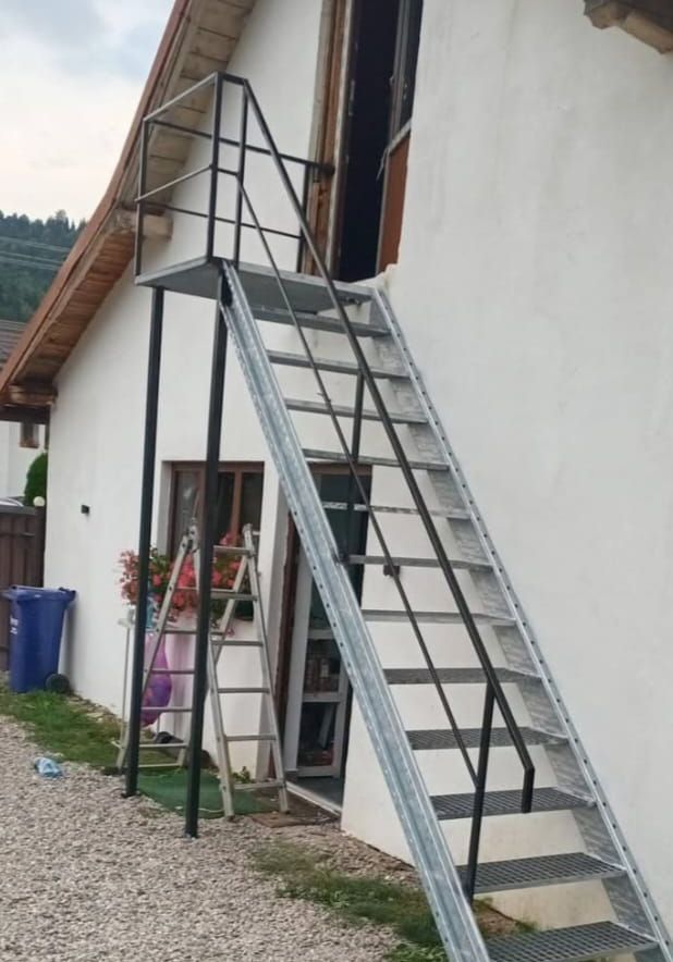 Scară exterioară zincată cu trepte din grătar, podest pe stâlpi negri și balustradă neagră, lângă peretele alb al unei case
